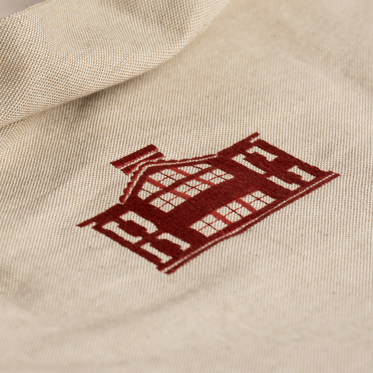 Close-up of a beige canvas or linen fabric with a dark red embroidered house logo.