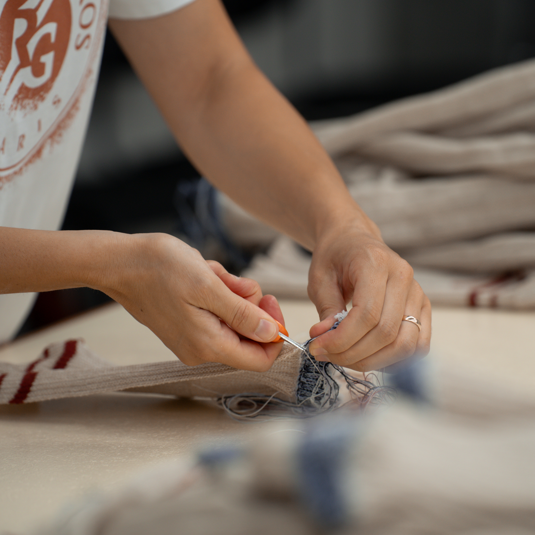 Close-up of a person's hands crocheting a beige knit garment with blue and red stripes using a crochet hook, with a soft-focus background of more fabric.