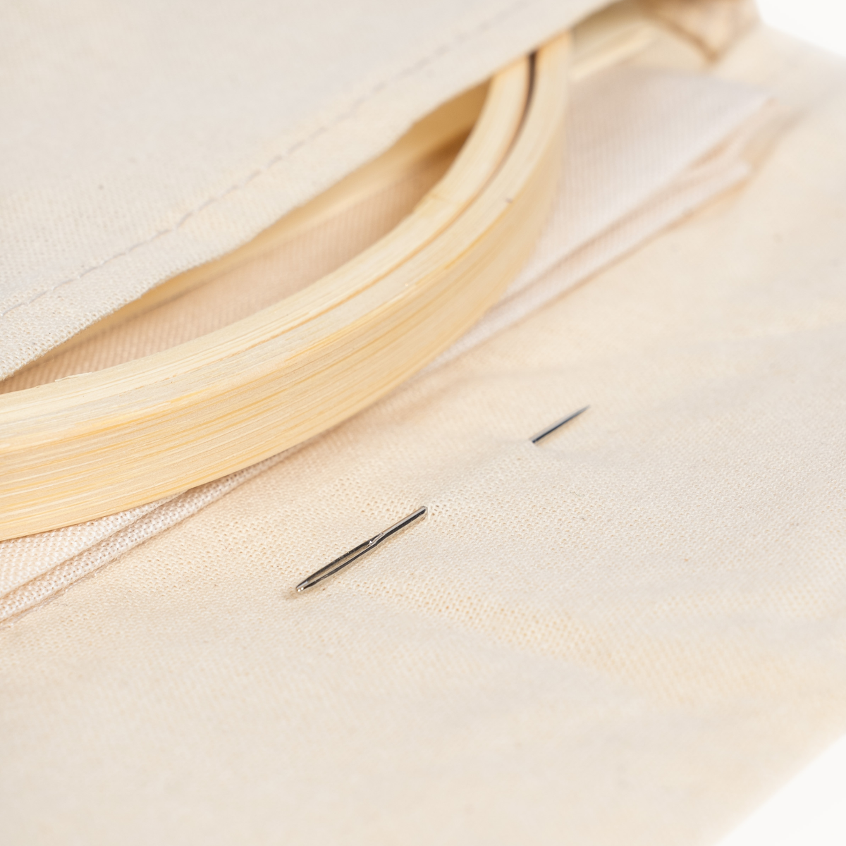 Close-up of a cream canvas embroidery hoop with a wooden frame and two metal embroidery needles on a matching canvas background.