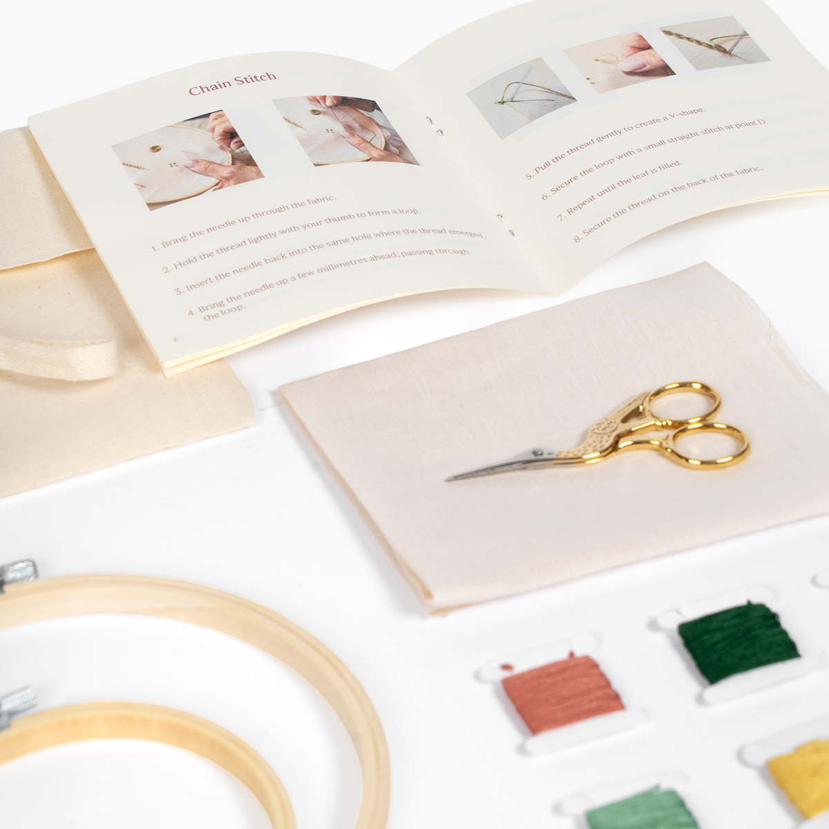 Embroidery kit laid out on a white surface, including a wooden hoop, thread spools in multiple colors, gold-handled scissors, a fabric pouch, and an open instruction booklet showing chain stitch steps.