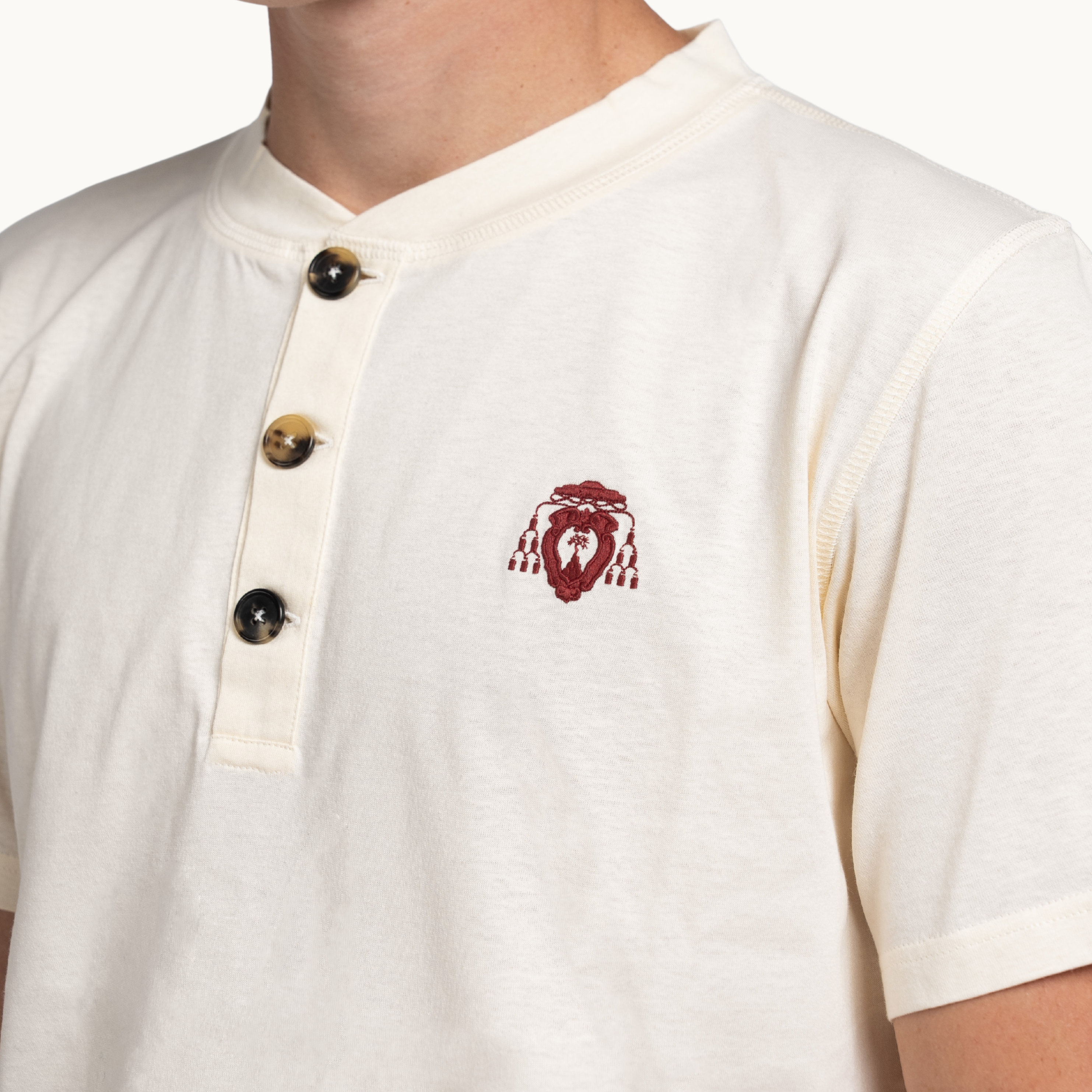 Close-up of a person wearing a cream-colored short-sleeve henley shirt with a red embroidered logo on the left chest and three dark buttons on the placket.