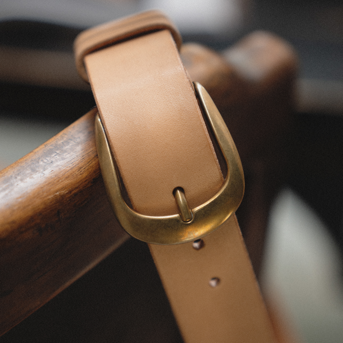 A close-up of a light tan leather belt with a polished brass buckle, draped over a dark wooden surface.