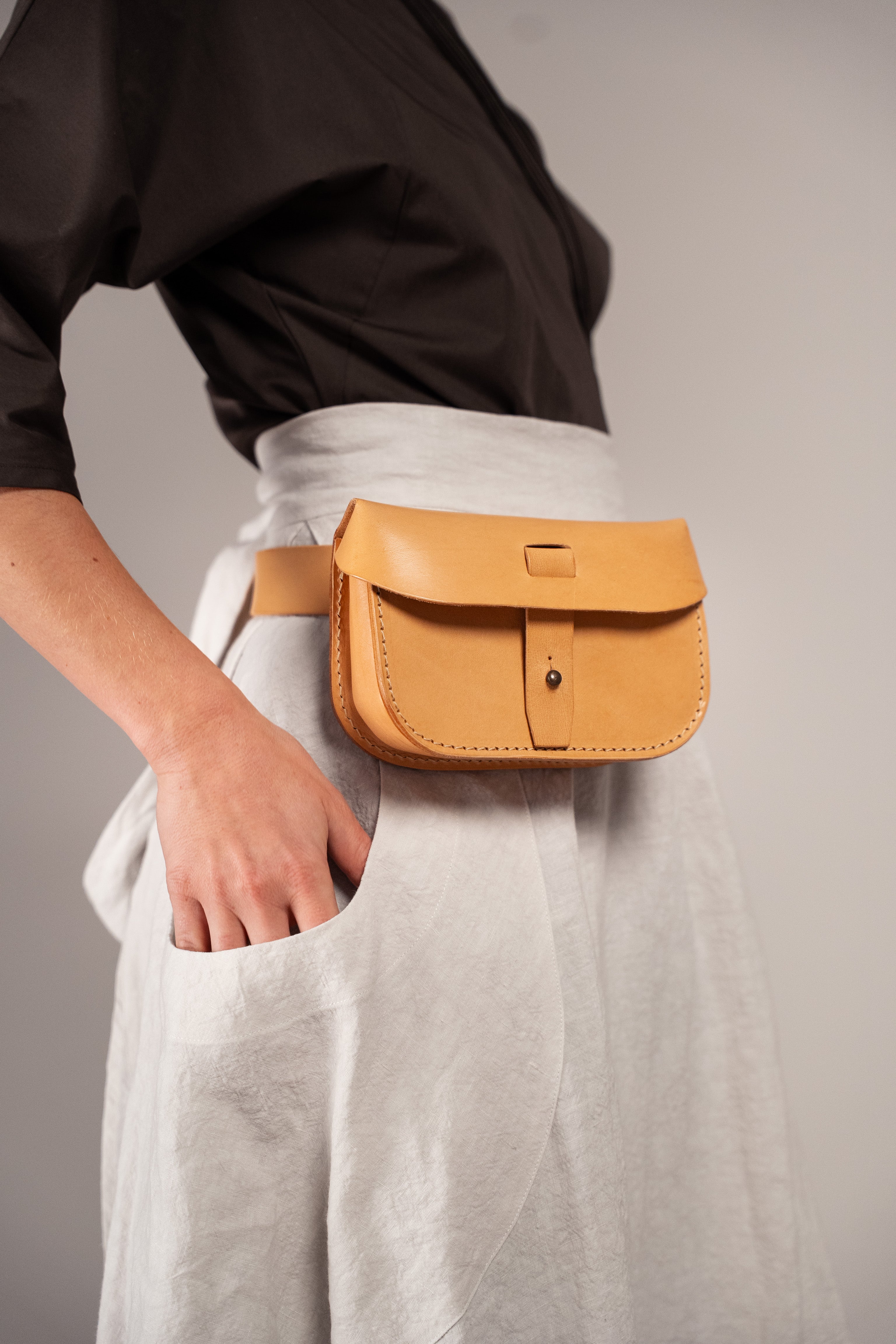 A tan leather belt bag with a front flap and metal snap closure, worn around the waist of a person in a black top and light gray skirt.