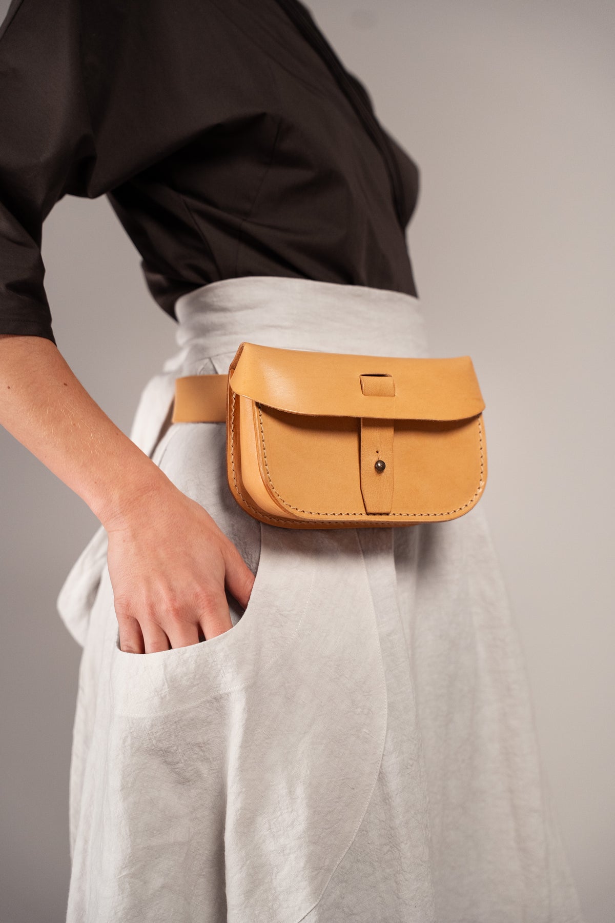 A tan leather belt bag with a front flap and metal snap closure, worn around the waist of a person in a black top and light gray skirt.