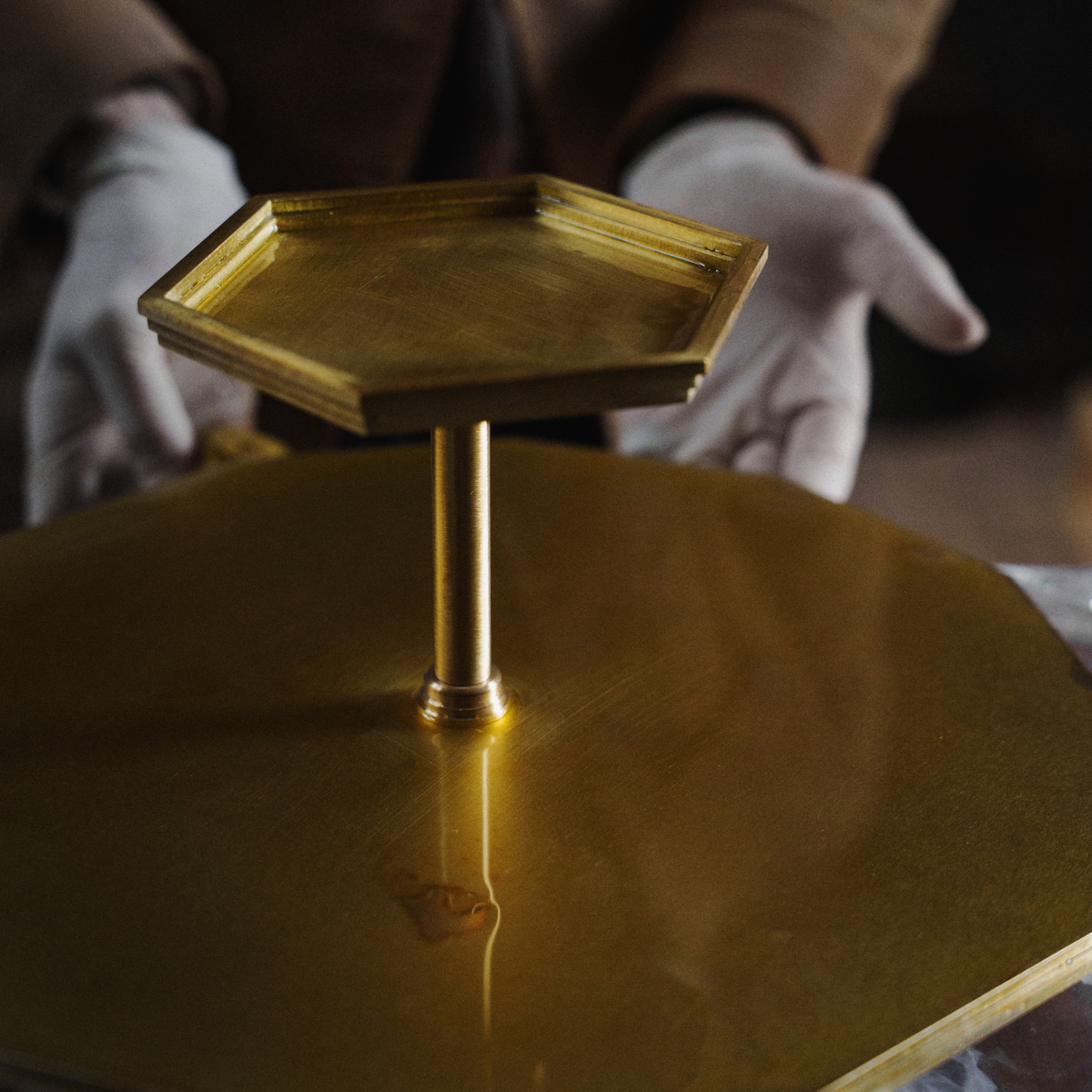 A brass two-tier serving stand with a hexagonal top tray and a central pedestal, shown with hands in white gloves holding it against a dark background.
