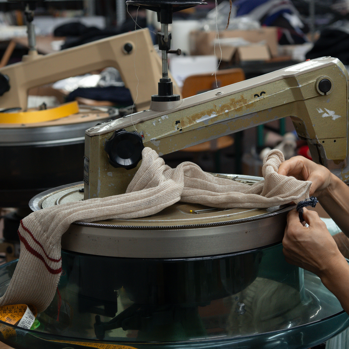 A close-up of a person's hands guiding a beige knit garment over a large industrial sewing machine in a workshop setting.