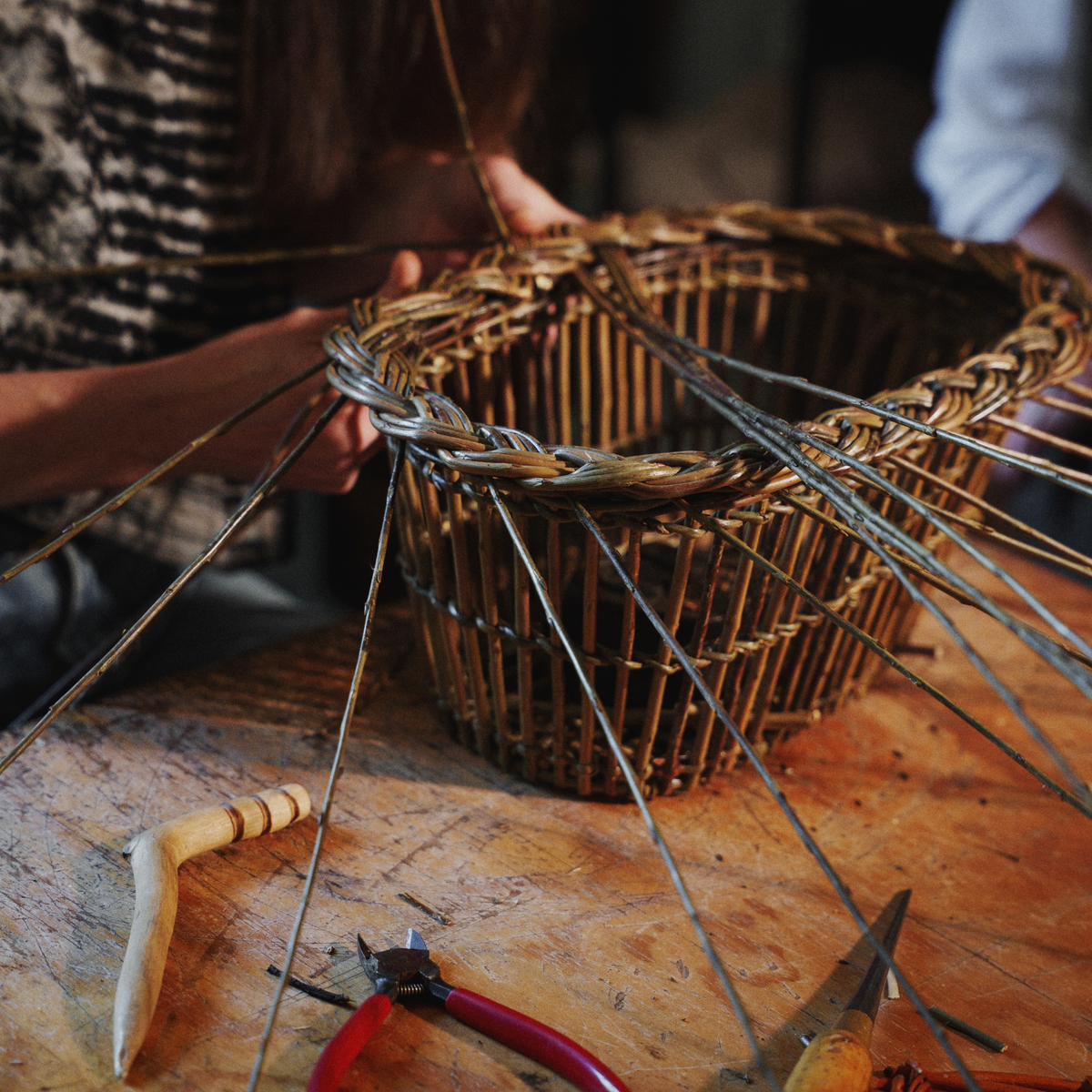 A close-up of a person weaving a round wicker basket on a wooden workbench, with thin string strands radiating outward and hand tools visible in the foreground.