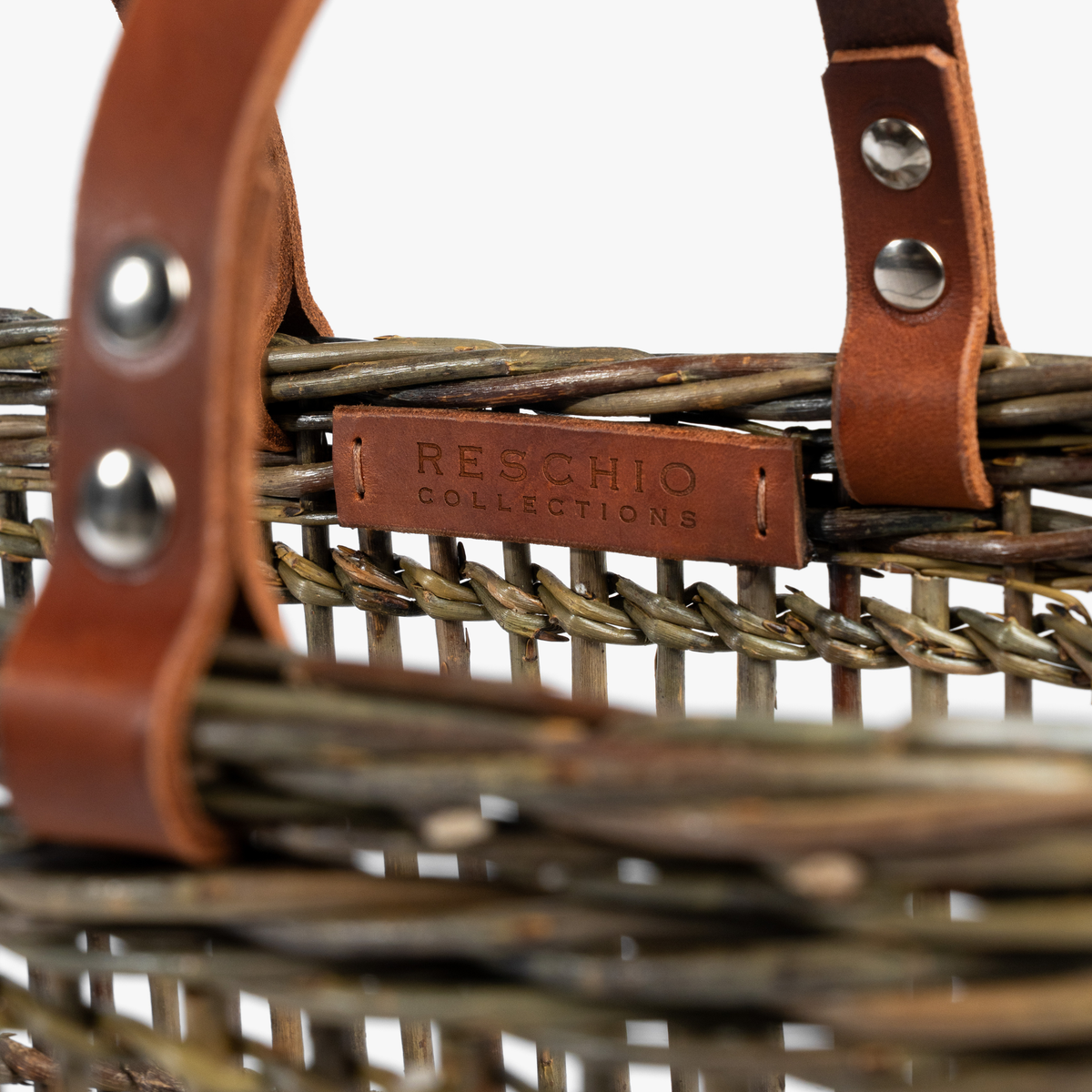 Close-up of a woven wicker basket with brown leather handles and a central embossed brand plaque reading 'RESCHIO COLLECTIONS' on a white background.
