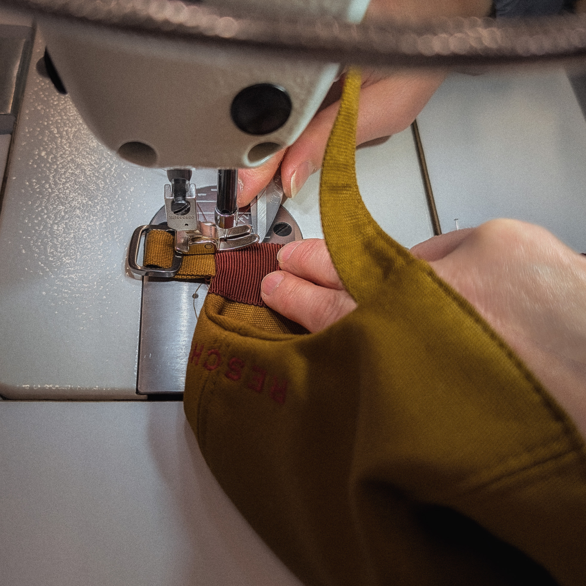 A close-up of a person's hands guiding a mustard yellow fabric face mask with a brown elastic strap under the needle of a sewing machine.
