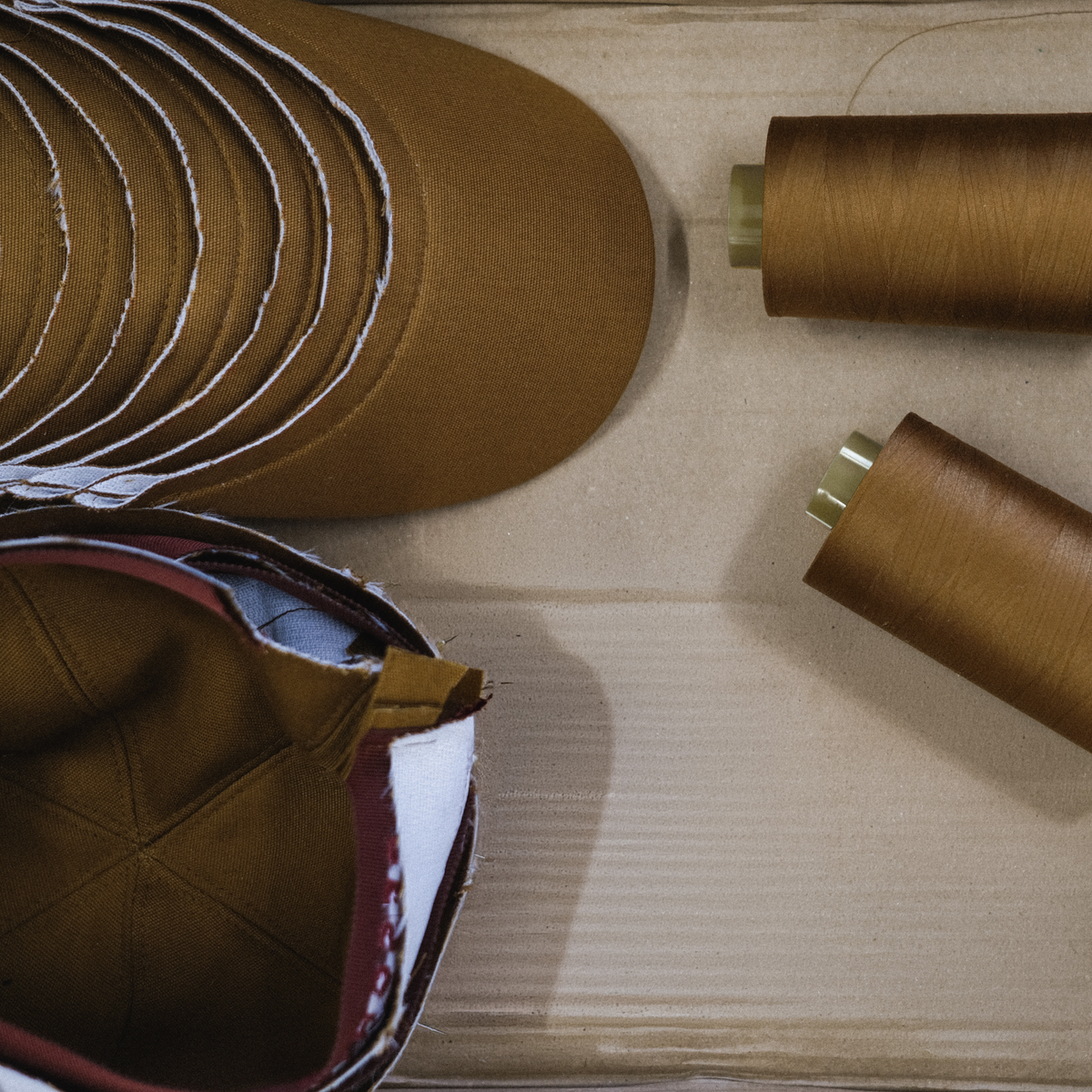 A close-up flat-lay of a brown fabric cap construction or sewing materials, including a partially assembled brown cap, stacked brown fabric pieces, and two spools of thread on a light wood surface.