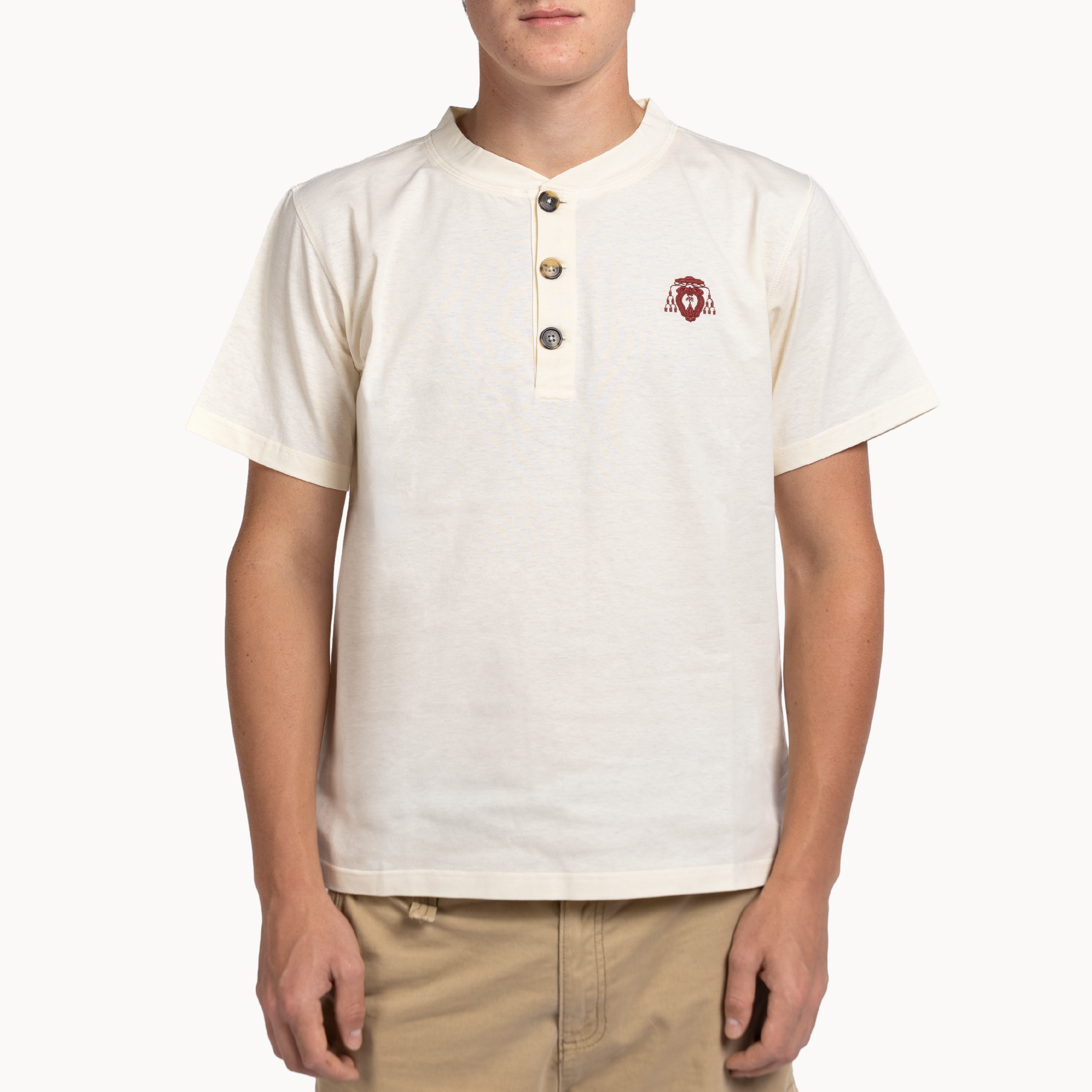 A man wearing a cream-colored short-sleeve henley shirt with a three-button placket and a small red embroidered logo on the left chest, paired with khaki pants, against a plain white background.