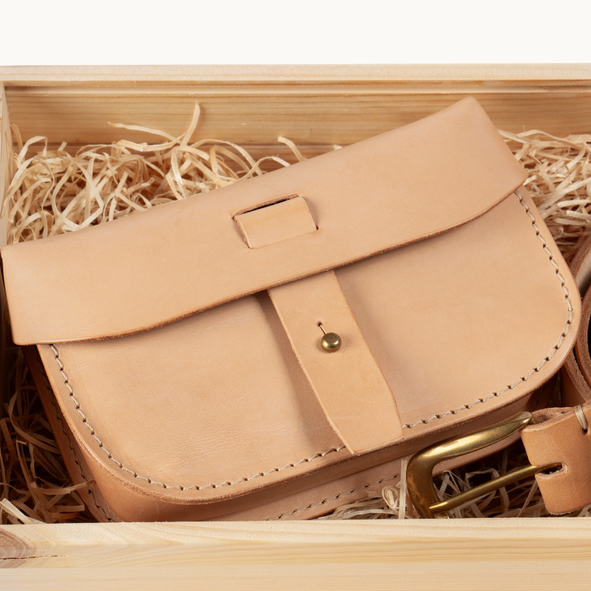 A light tan leather saddle-style pouch with a flap closure and brass snap button, nestled in shredded paper inside a wooden box.