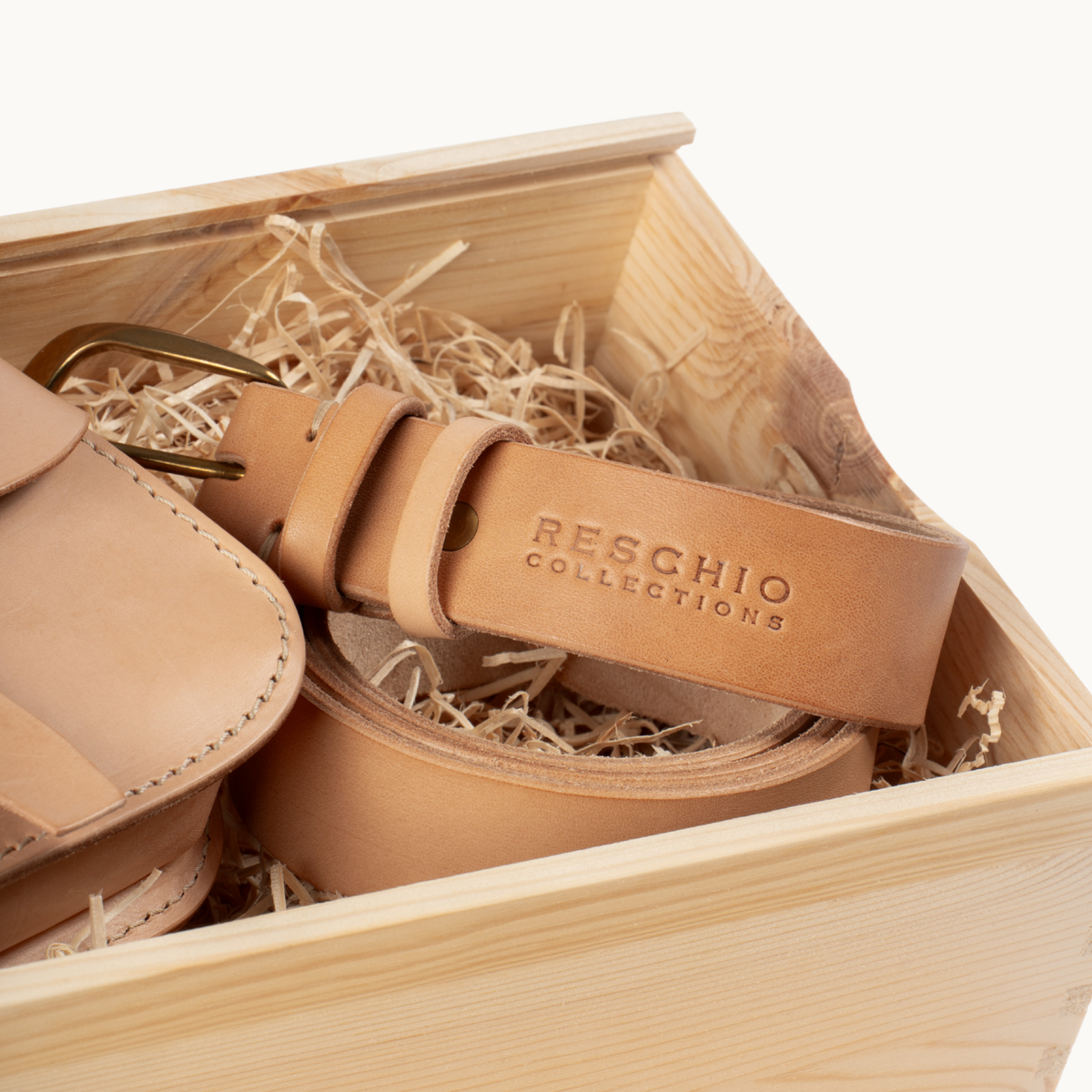 A tan leather belt with a brass buckle and a matching leather pouch, presented inside a light wood gift box filled with shredded paper. The belt has 'RESCHIO COLLECTIONS' embossed on the keeper.