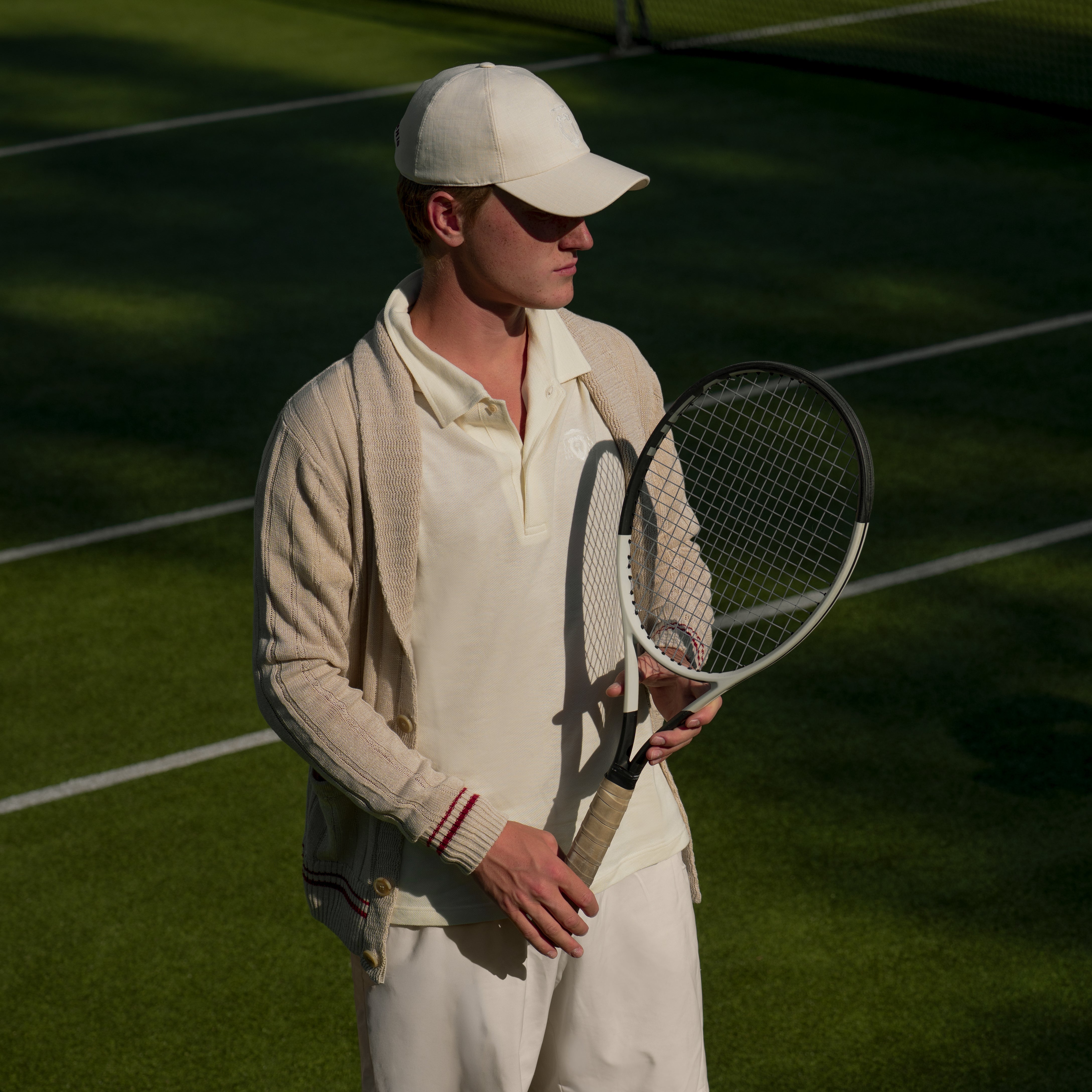 A man wearing a cream-colored tennis uniform, including a cap, polo shirt, sweater, and trousers, stands on a grass tennis court holding a tennis racket.