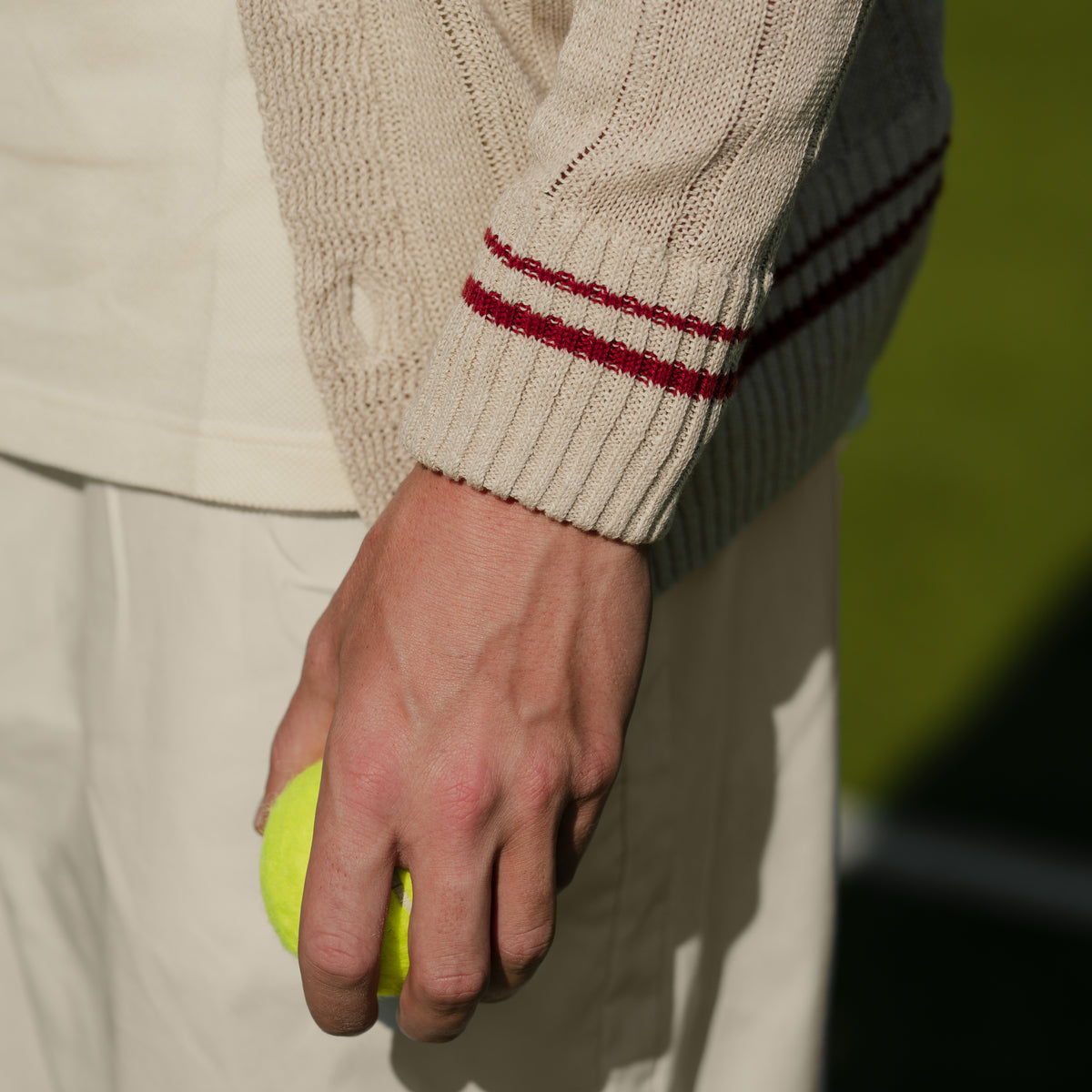 Close-up of a person holding a yellow tennis ball, wearing a cream knit sweater with red stripes on the cuff and cream trousers, against a blurred green background.