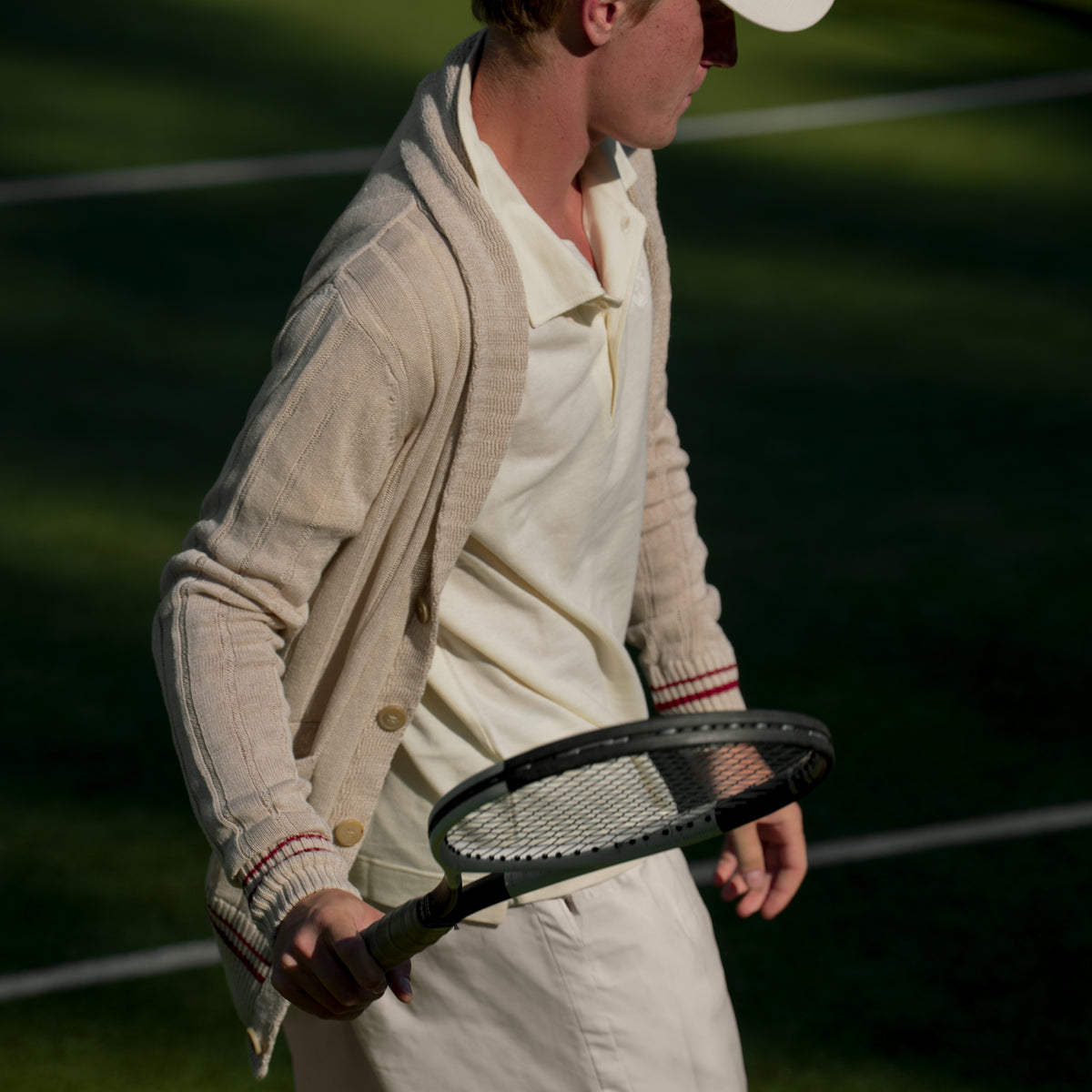 A man on a tennis court wearing a cream-colored cricket uniform with a beige knit cardigan, holding a vintage tennis racket.