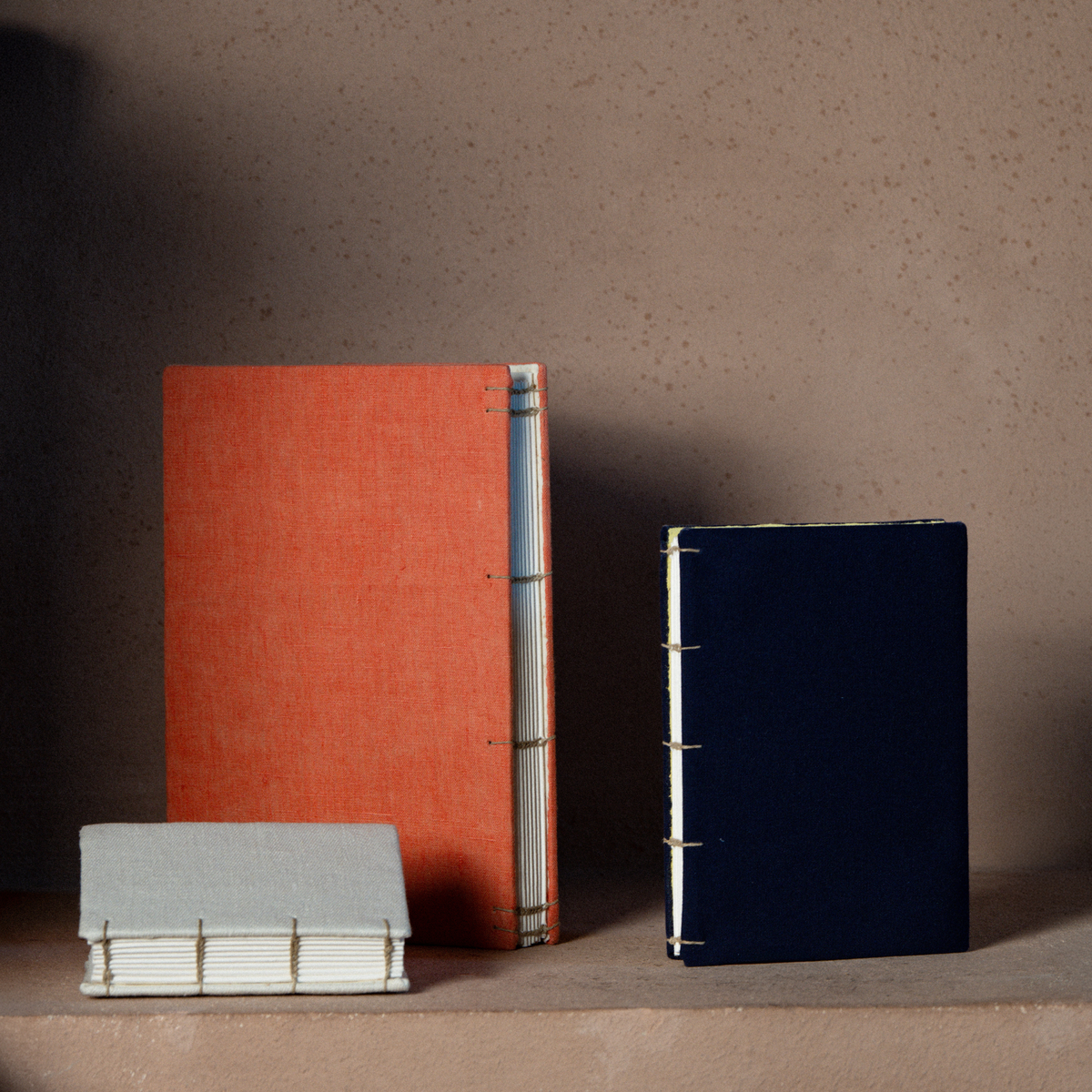 Three handmade bound books or notebooks displayed on a shelf against a textured beige wall. A small light gray book lies in front, a tall orange book stands in the back left, and a dark navy book stands in the back right.
