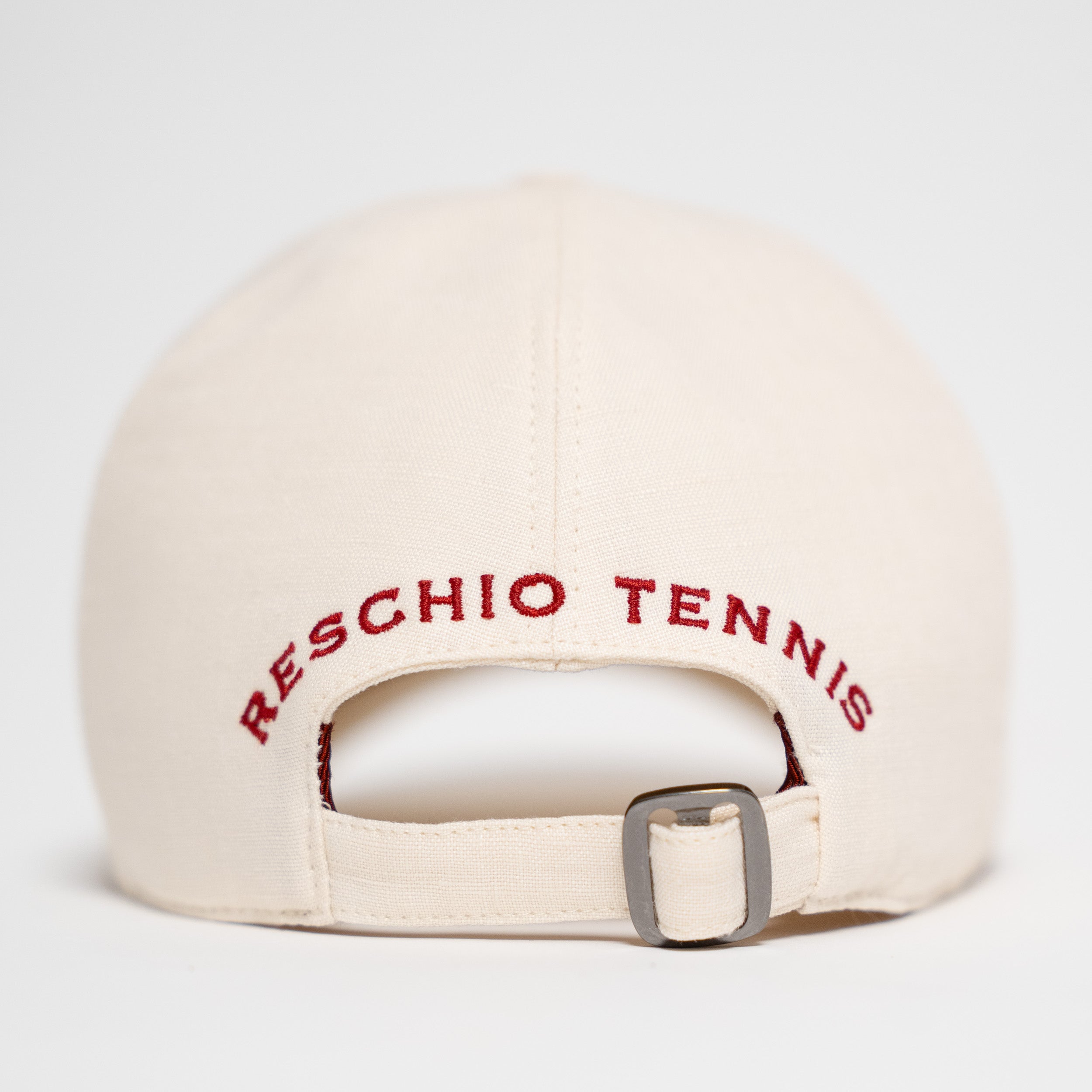 Back view of an off-white baseball cap with 'RESCHIO TENNIS' embroidered in red across the back strap, featuring a silver metal buckle.