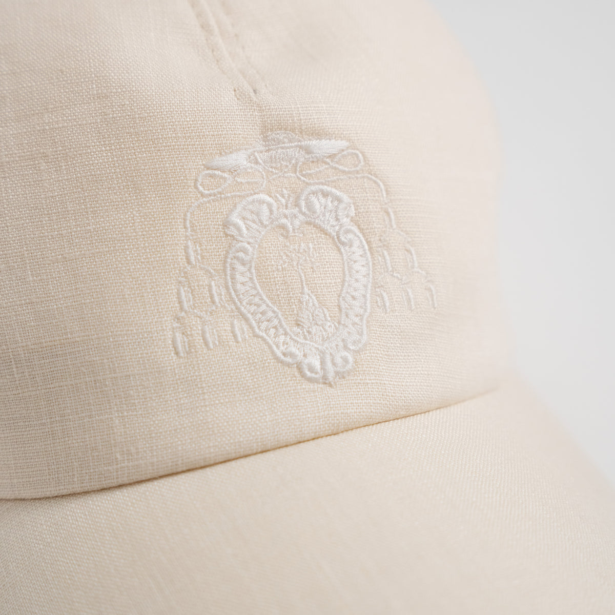 Close-up of a light cream or off-white textured fabric cap with an embroidered white crest logo on the front panel.