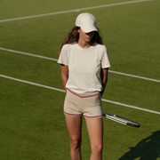 A woman stands on a grass tennis court wearing a white t-shirt, cream-colored tennis shorts with red stripes, and a white baseball cap, holding a tennis racket.