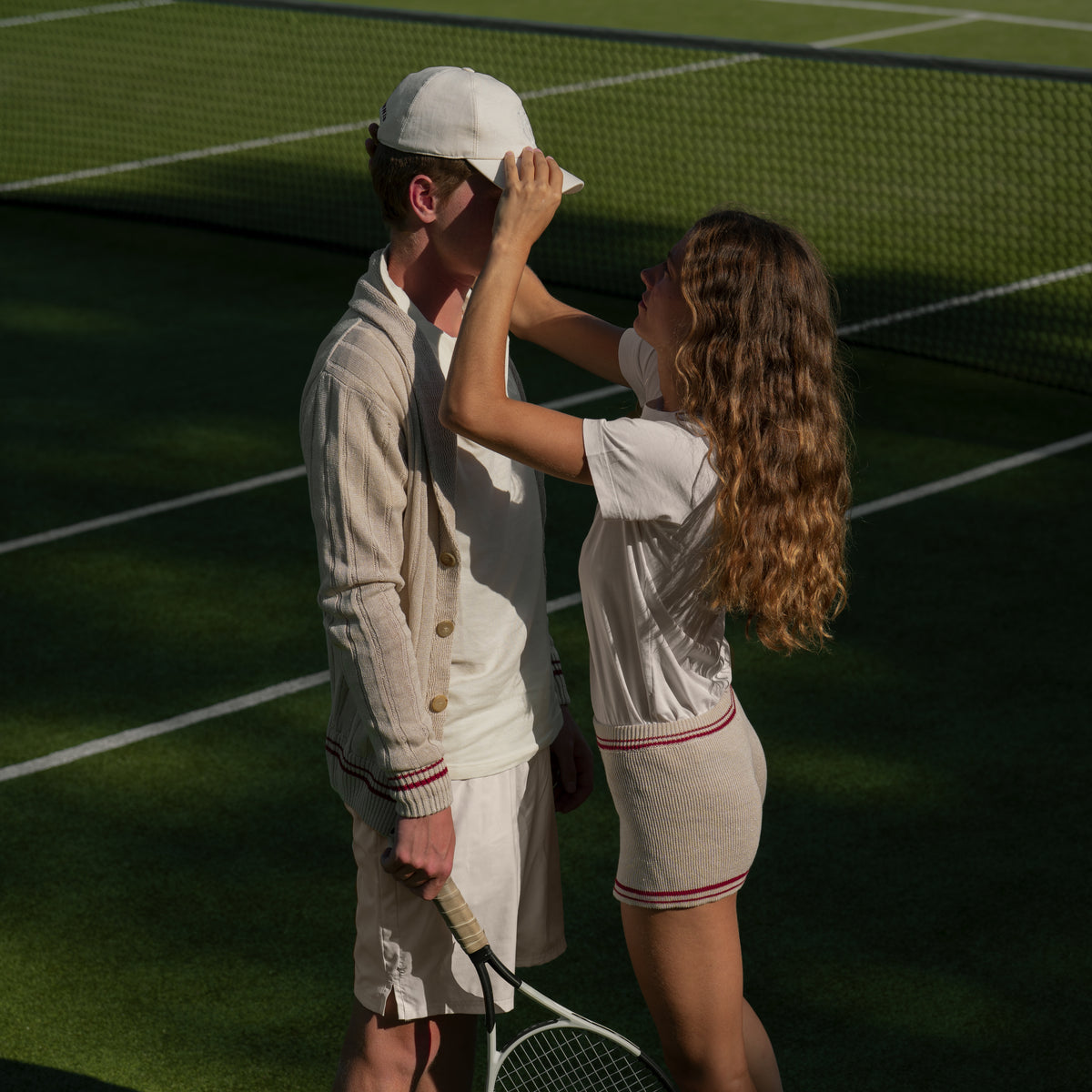 Two people in coordinated cream tennis outfits standing on a green tennis court, with one person adjusting the other's cap. A tennis racket is visible.