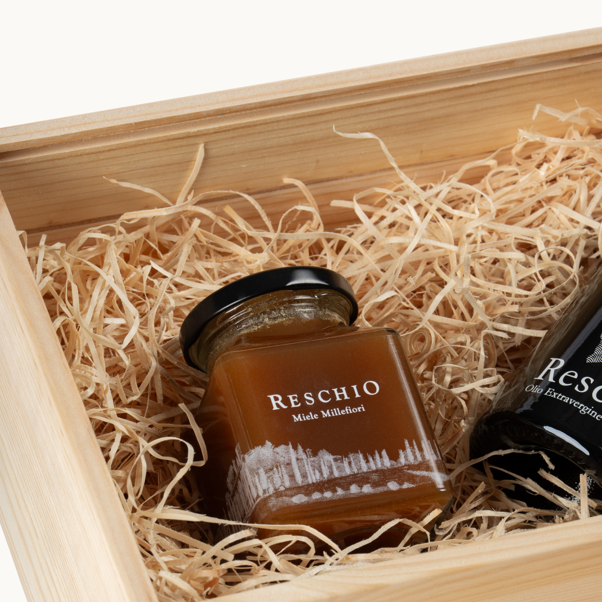 A glass jar of Reschio Made Milfiori honey nestled in light tan paper shred inside a wooden gift box, with part of a dark bottle visible beside it.