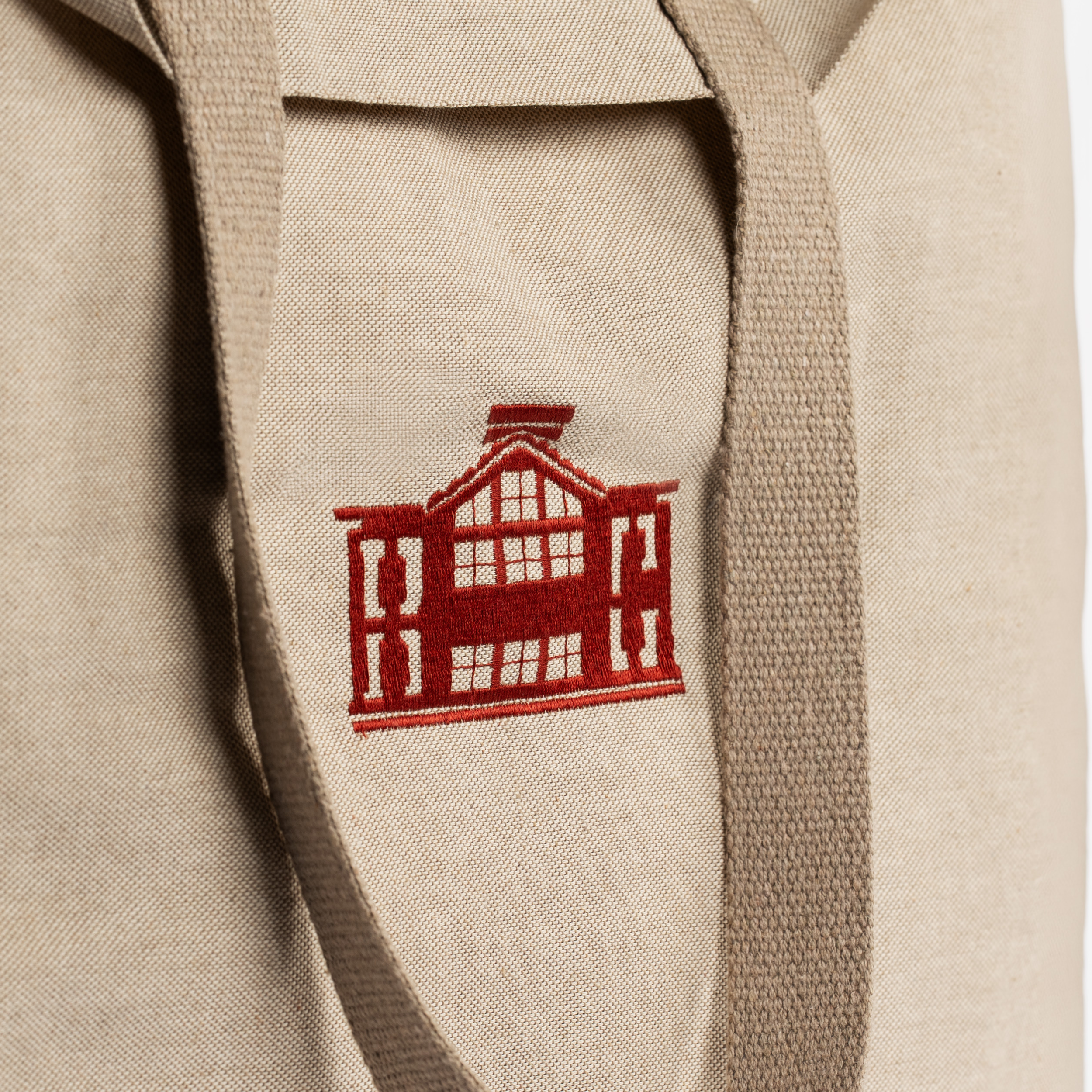 Close-up of a beige canvas tote bag with a red embroidered house logo on the front and matching beige shoulder straps.