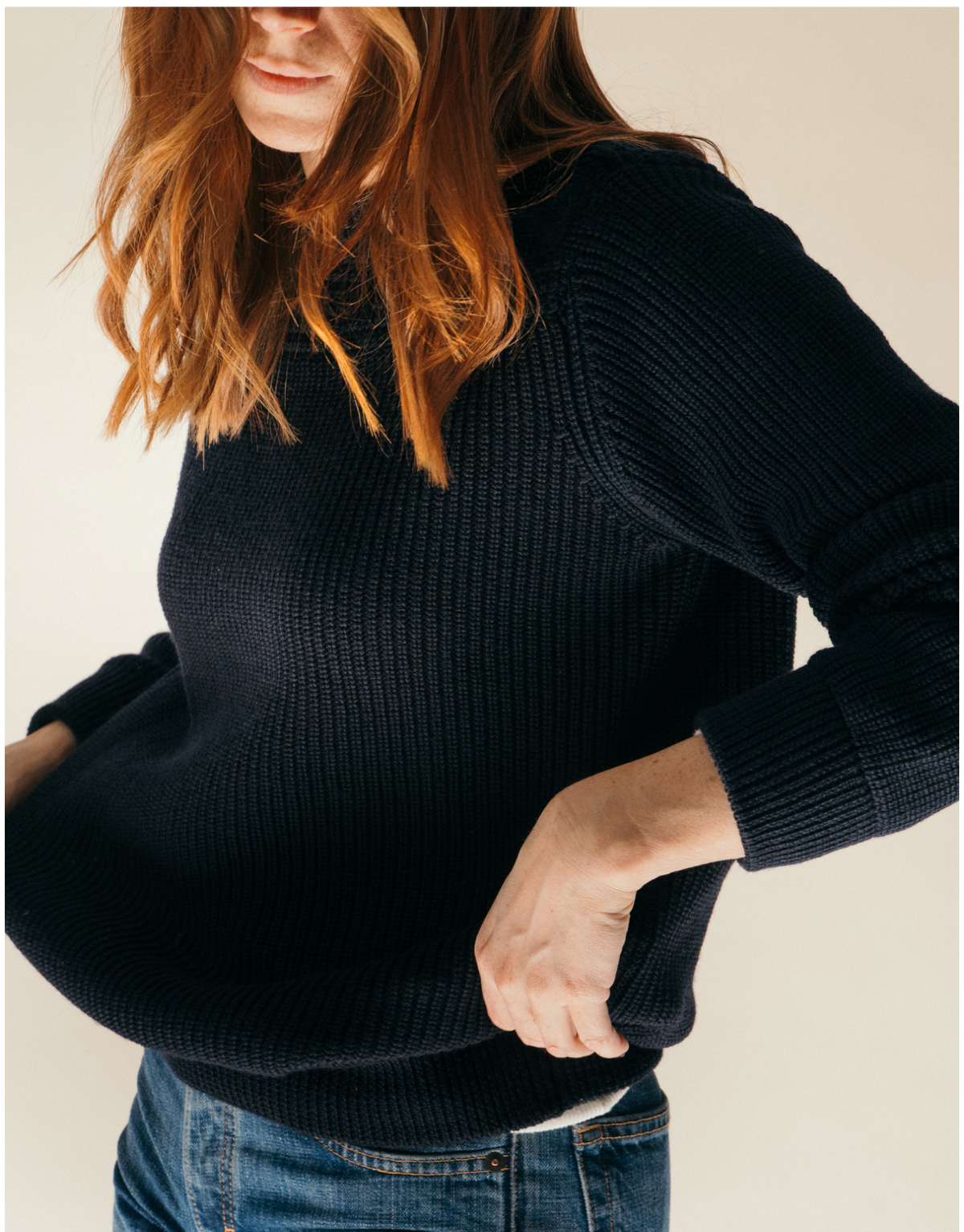 A woman wearing a dark navy ribbed knit sweater and blue jeans, shown from the chest down against a plain light beige background.