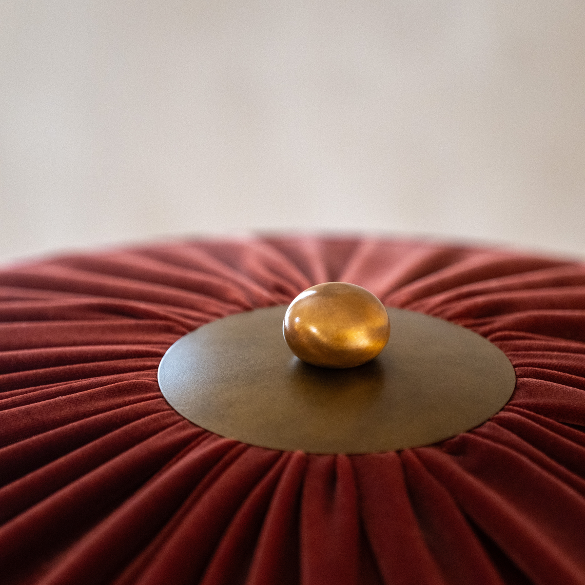 A close-up of a round red velvet pouf or ottoman with pleated radial texture, a central metallic gold base, and a small polished gold knob on top.