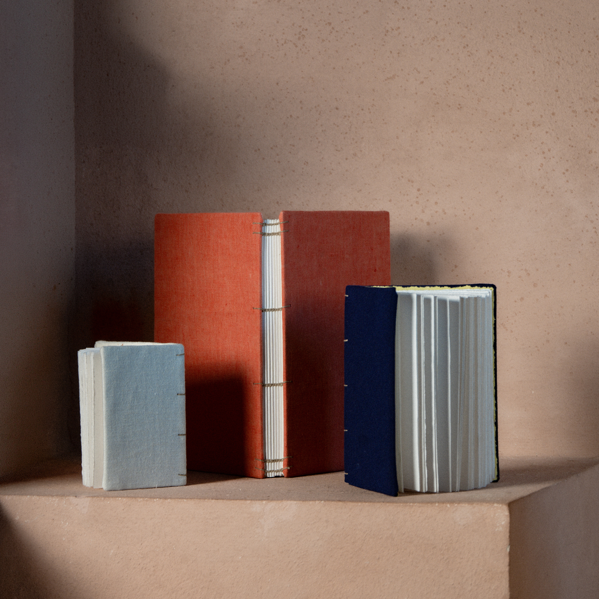 Three handmade paper books with colored covers standing upright on a matching beige pedestal against a textured beige wall.