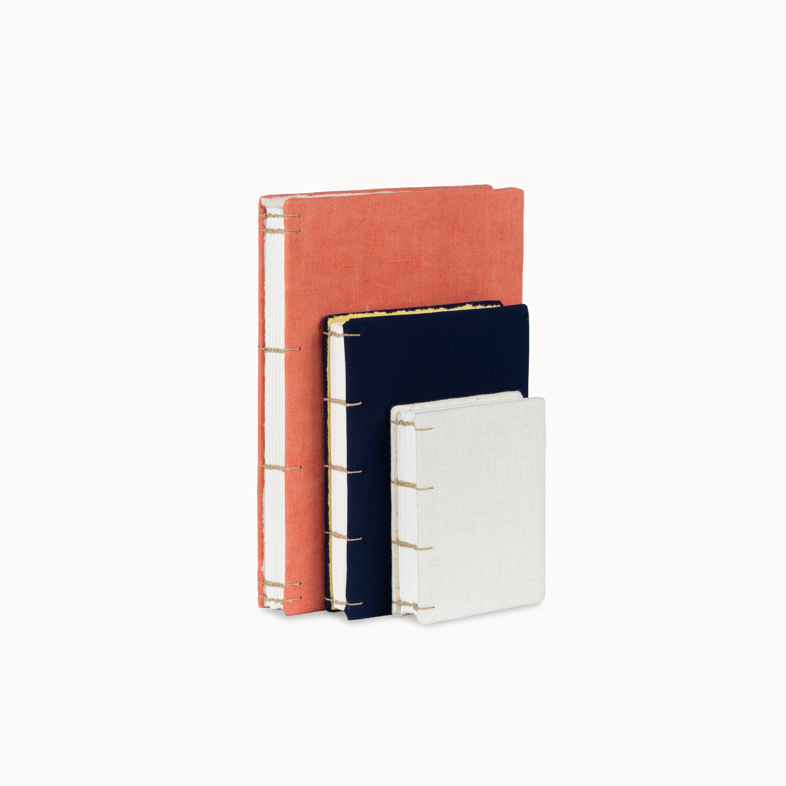 Three linen-bound notebooks in coral, navy, and white, arranged in a vertical stack with gold-tone wire binding on a clean white background.