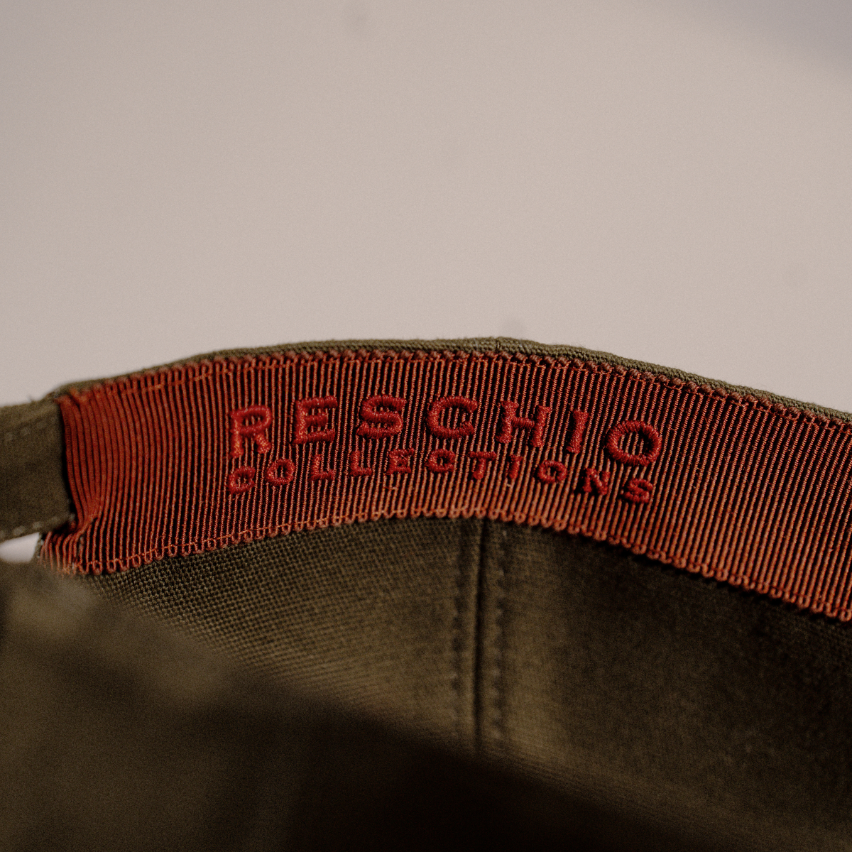 Close-up of the interior collar of an olive green cap, showing a red-orange grosgrain ribbon label with the brand name 'RESCHIO COLLECTIONS' embroidered in red.